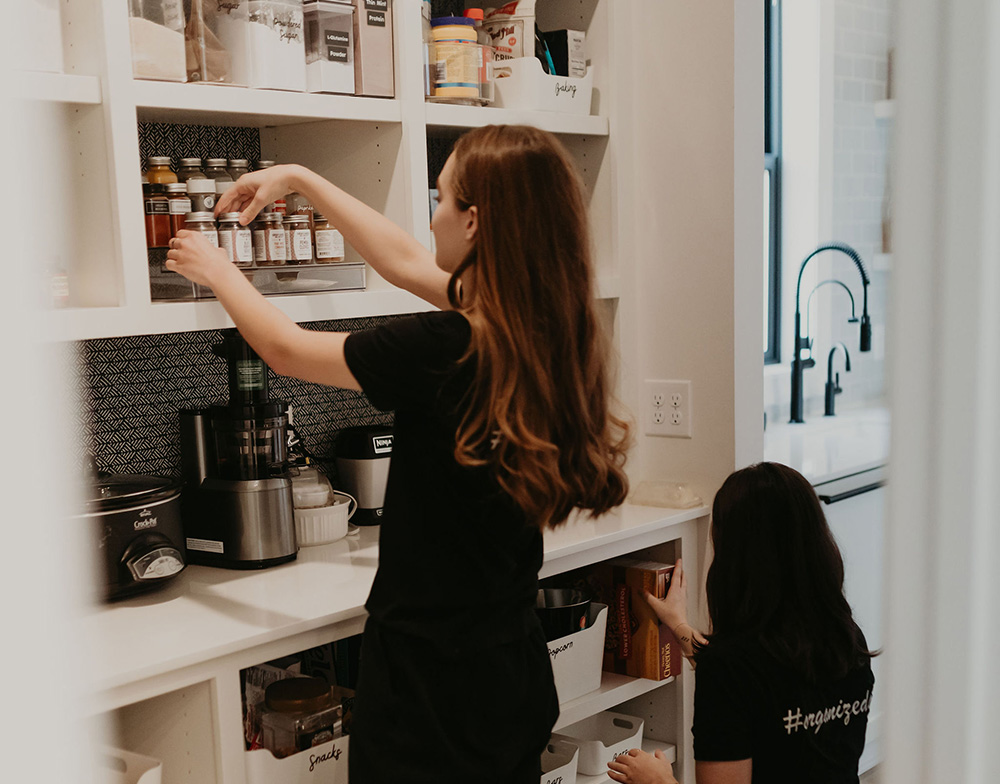 A team of professional organizers from Meat and Potatoes Organizing arranging items in a neatly organized pantry in a home. Specializing in pantry organization, kitchen decluttering, and efficient storage solutions in Minneapolis and Saint Paul.